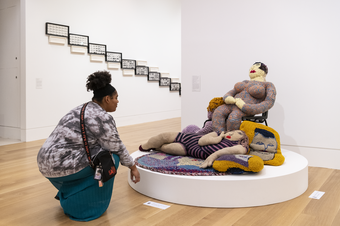A visitor to the gallery crouches down to look at a large fabric sculpture of several figures on a low circular plinth